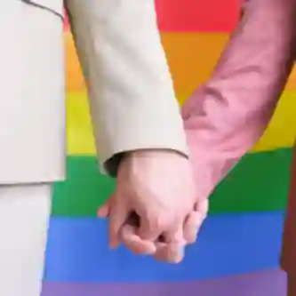 Splecione dłonie na tle kolorowej tęczowej flagi LGBTQ+.