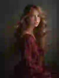 Side view of a woman with wavy hair in dark red ruffled dress sitting and staring at the camera. Her hair flows because of the breeze. Template for Undress AI.