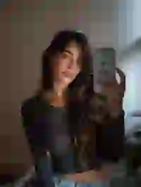 Portrait of a woman in black long sleeve crop top taking selfie with a white iPhone in a bedroom. Template for Undress AI.