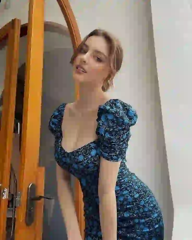 Eine Frau in einem engen blauen Blumenkleid beugt sich an einer Tür vor und zeigt, wie man sie auszieht.