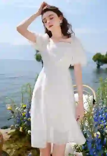 A woman with dark wavy hair in a white dress is posing beside a lake with flowers in the background.