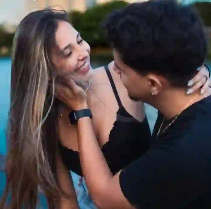 Photo of a loving couple. The woman, wearing a black bra, smiles affectionately at her partner with her hand resting on his shoulder. Only the back of the man is visible, with one of his hands gently cupping her cheek.